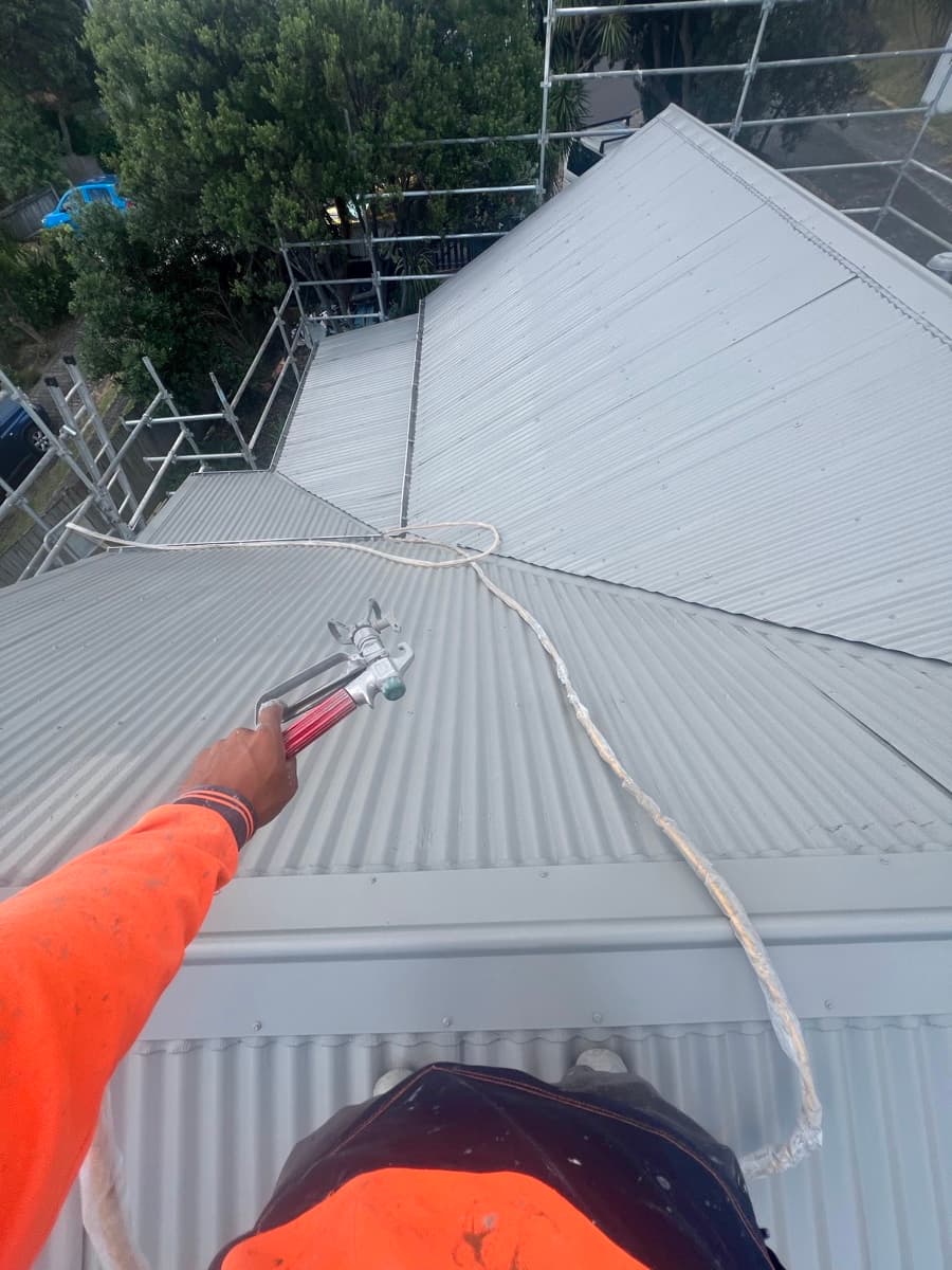 Roofer in orange hi-vis holding a spray gun, respraying a corrugated iron roof from scaffolding